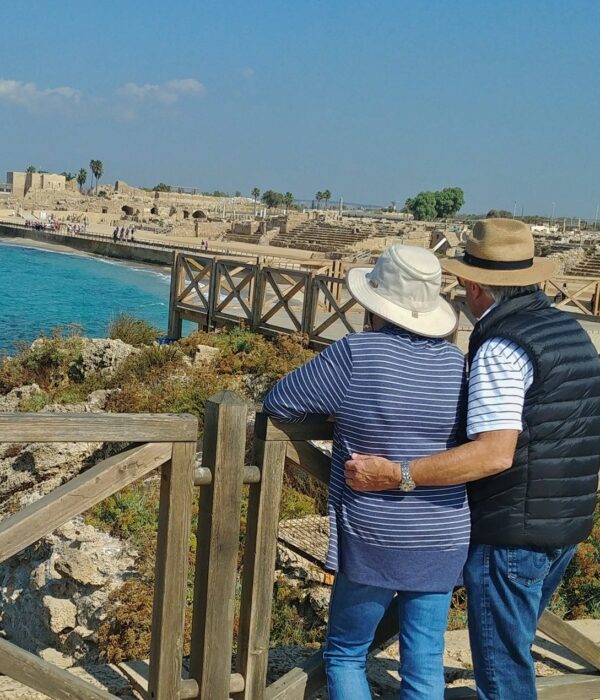 A couple in sun hats stand arm in arm at a wooden railing, looking over the turquoise sea and the stone seating of the ancient hippodrome at Caesarea.