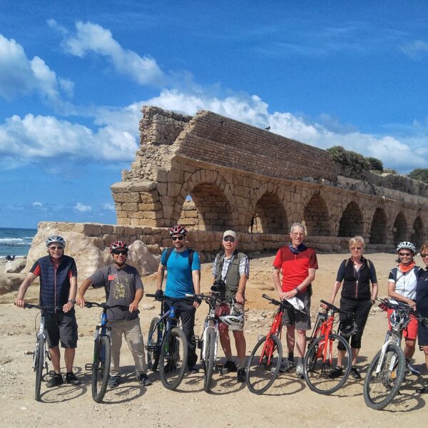 A group of cyclists with bikes on the beach in front of the Roman aqueduct at Caesarea
