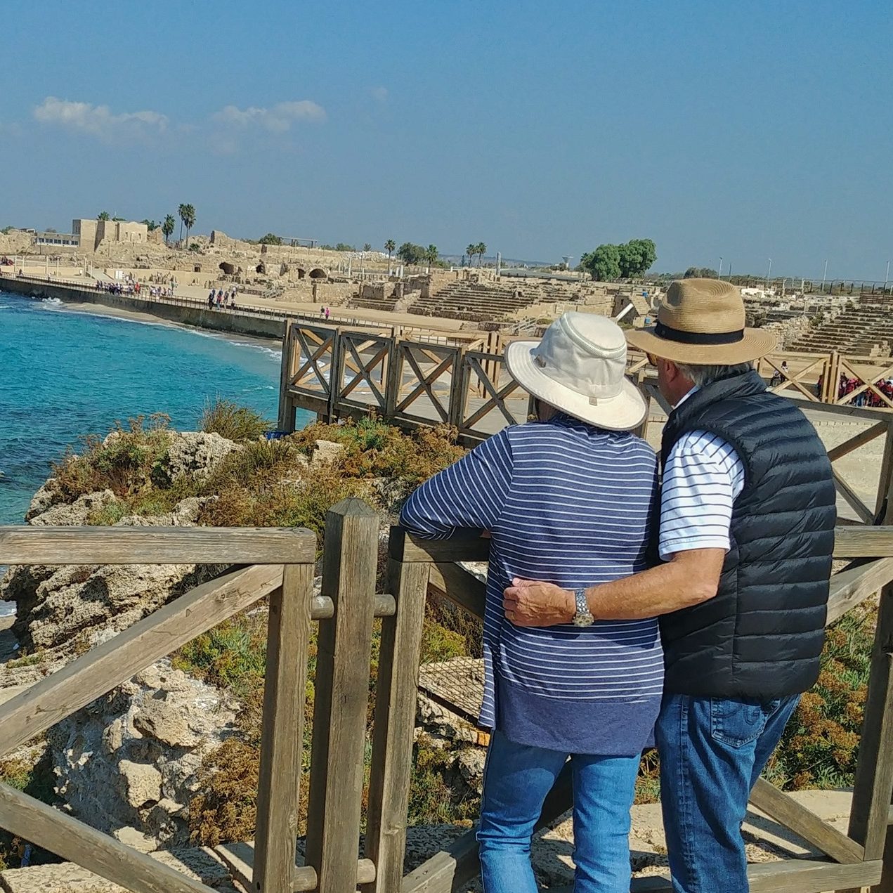 Caesarea tour: Herod’s hippodrome by the Mediterranean A couple in sun hats stand arm in arm at a wooden railing, looking over the turquoise sea and the stone seating of the ancient hippodrome at Caesarea.