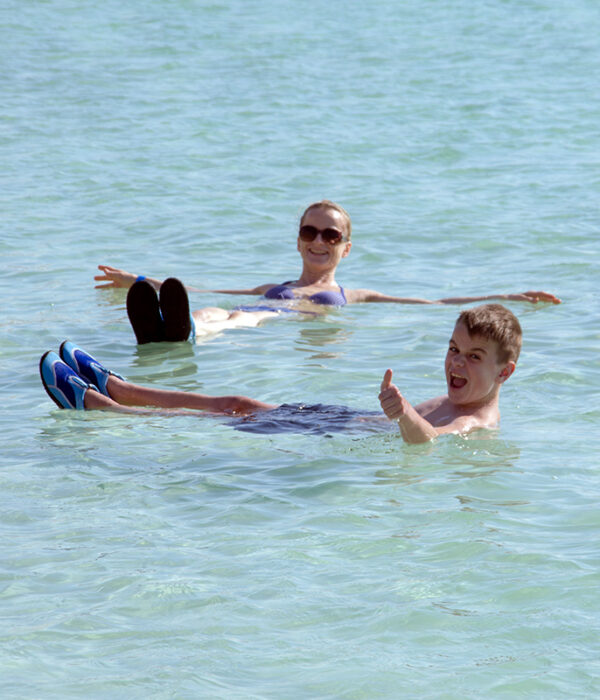 A smiling woman and a boy giving a thumbs-up float on their backs in the clear, calm water of the Dead Sea.