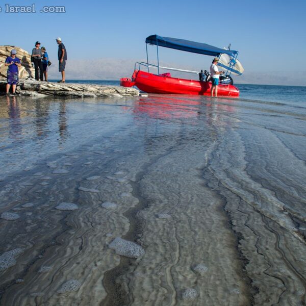 A small group stands on salt rocks by a red boat on the Dead Sea