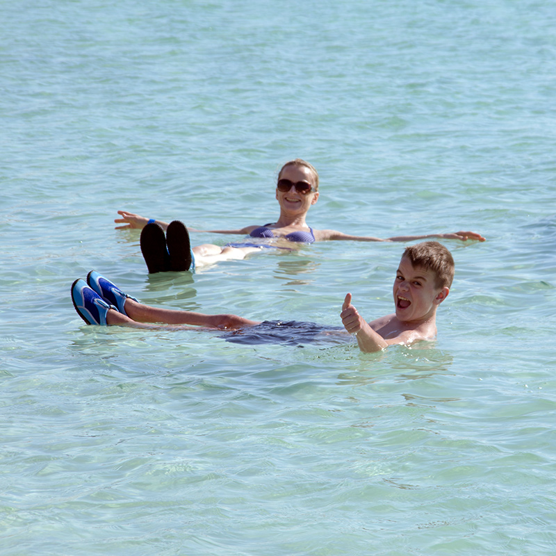 Dead Sea tour: floating at the lowest point on Earth A smiling woman and a boy giving a thumbs-up float on their backs in the clear, calm water of the Dead Sea.