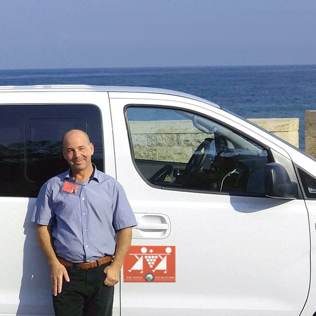 Tour guide Udi Avital standing beside his white tour van, with the sea behind a stone wall, Israel.