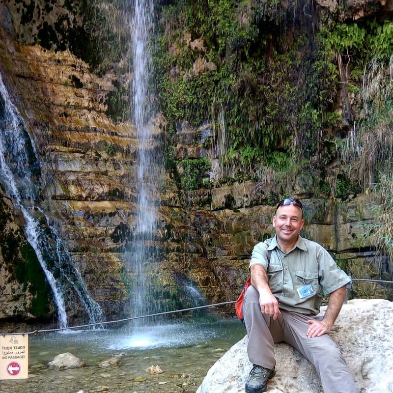 Ein Gedi tour: David’s Waterfall with guide Udi Avital Tour guide Udi Avital smiles, sitting on a boulder beside David's Waterfall in Ein Gedi, where a tall thin waterfall drops down a green, fern-covered cliff into a pool.