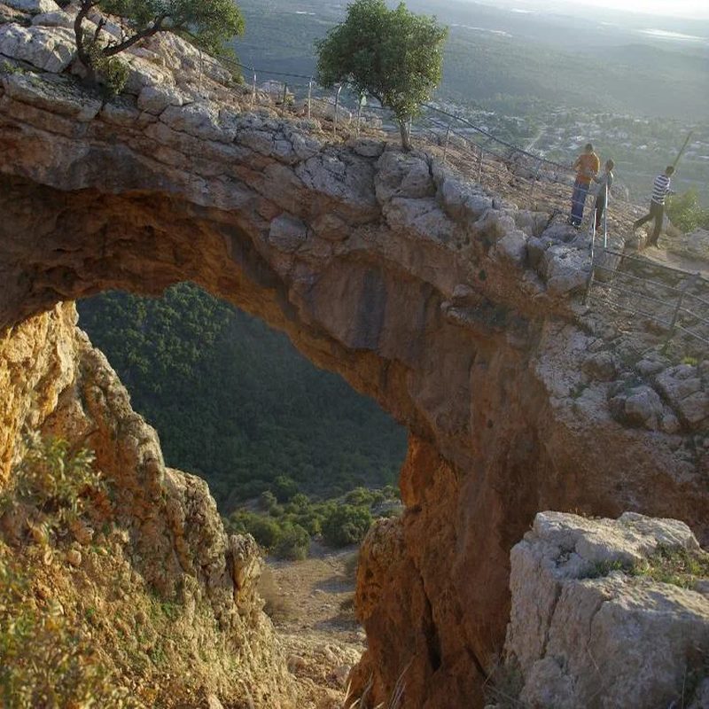 Galilee tour: the Keshet Cave rock arch above the green hills A huge natural limestone arch at Keshet Cave frames a forested valley in the Galilee, with visitors standing at a railing on the clifftop above.